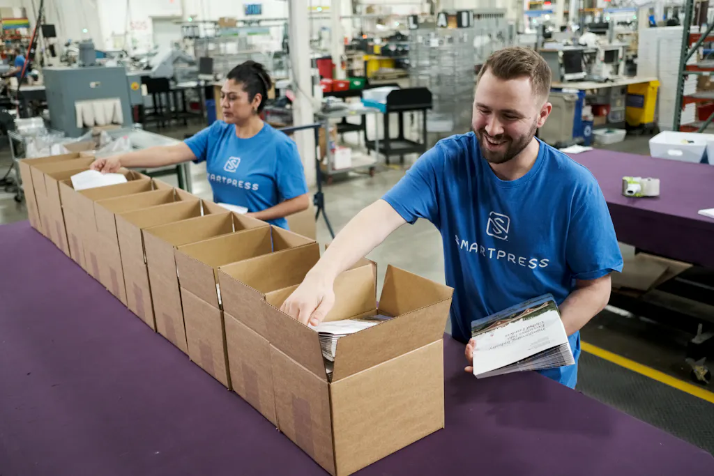 A woman and a man wearing Smartpress T-shirts packaging products into cardboard boxes in a production facility.