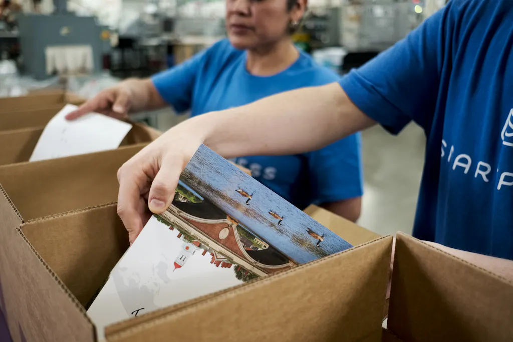 A woman and a man wearing Smartpress T-shirts packing print products in cardboard boxes in a production facility.