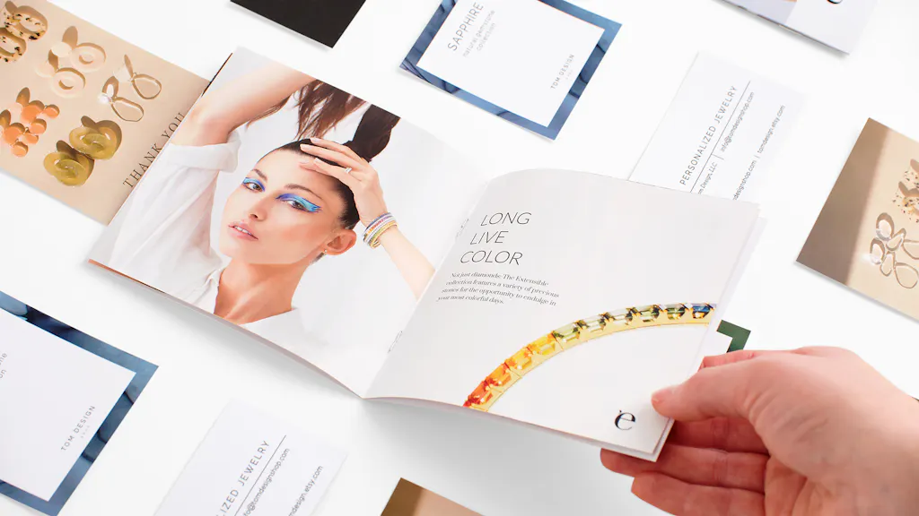 A hand holding open a jewelry product booklet with other jewelry print assets around it.