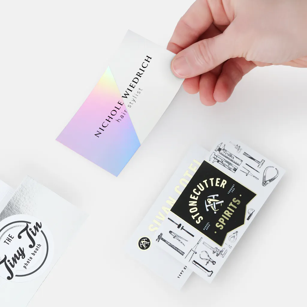 A hand holding a holographic foil business card with silver and gold foil business cards around it.
