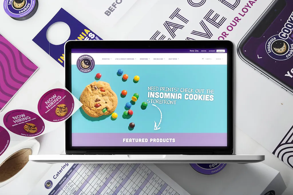 A laptop showing the Smartpress Storefront for Insomnia Cookies with branded print marketing behind it.