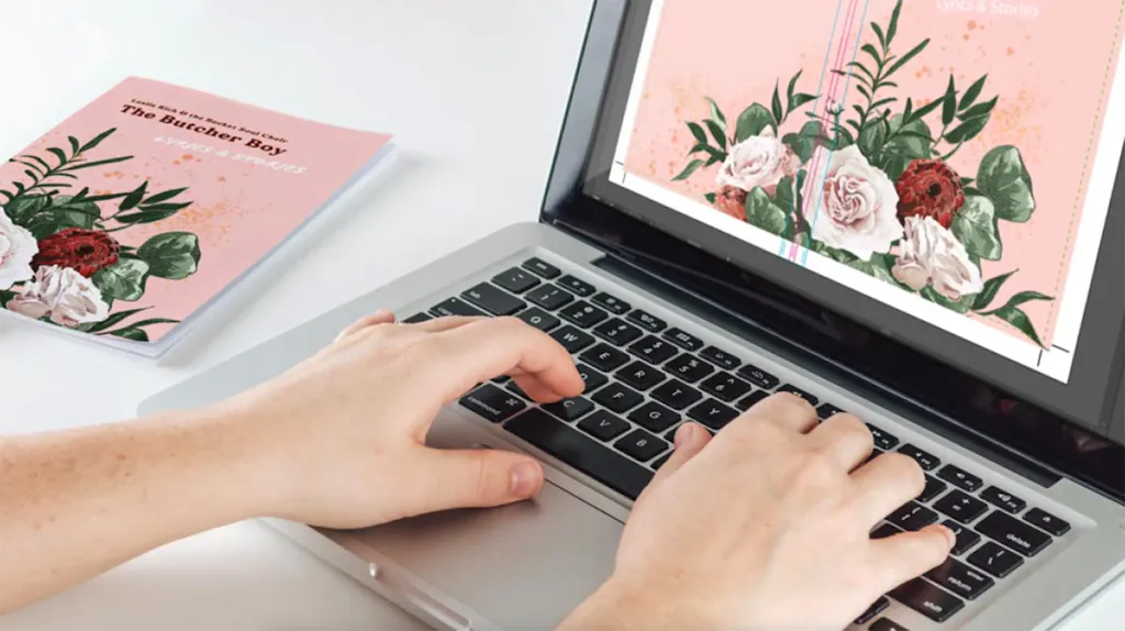 Two hands on a laptop designing a song and lyrics booklet with a pink and floral design.