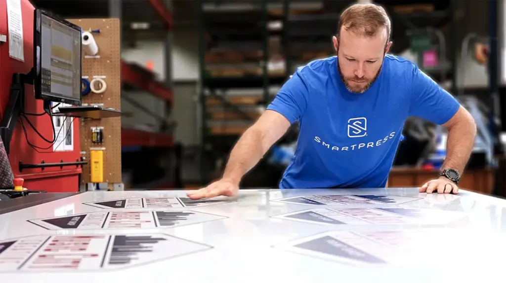 A man in a blue Smartpress T-shirt leaning over a production table with custom aluminum signs on it.