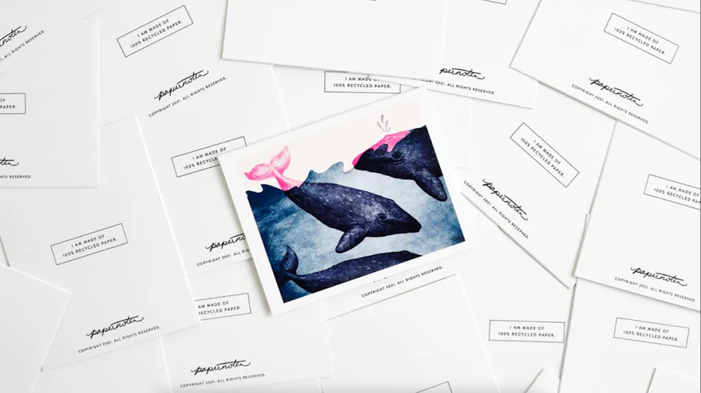 A postcard printed with whale artwork laying on top of a pile of scattered postcards with I am made of recycled paper.